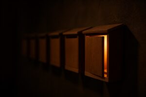 暗い壁に並ぶ木製の郵便受け。1つだけ扉が開いて中から暖色の光が漏れている。タグではなく個人の入口から入るという記事の比喩